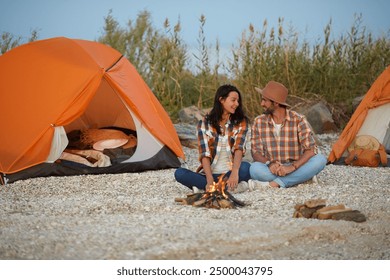 A couple sitting together by a campfire at their beachside campsite with orange tents, sharing a moment of warmth and companionship. - Powered by Shutterstock