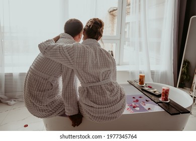 the couple is sitting in white lab coats with their backs to the camera, looking out the window while sitting on the bathroom. There are rose petals and candles all around. Romantic setting - Powered by Shutterstock