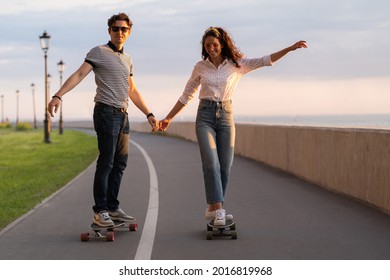 Couple of skateboarder riding skates holding hands. Happy young adults enjoy longboarding together on sunset on pavement near seaside. Casual stylish man and woman in trendy outfit skating outdoors - Powered by Shutterstock