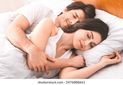 A couple sleeps peacefully in bed together at home. The woman is lying on her side, her head resting on a pillow, while the man is hugging her from behind. They both have their eyes closed - Powered by Shutterstock