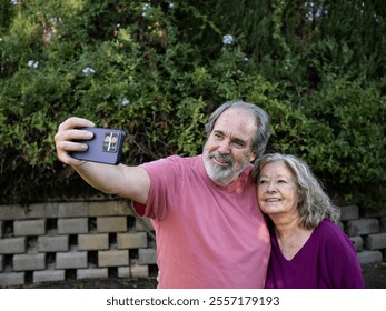 A couple smiles happily while taking a selfie in a lush garden. They are surrounded by greenery and blooming flowers, enjoying a sunny morning together. - Powered by Shutterstock