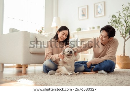 Couple smiles while playing with their dog on a soft rug in a well-lit living room.
