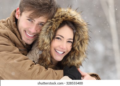Couple smiling with perfect teeth hugging and looking at camera in winter in a forest