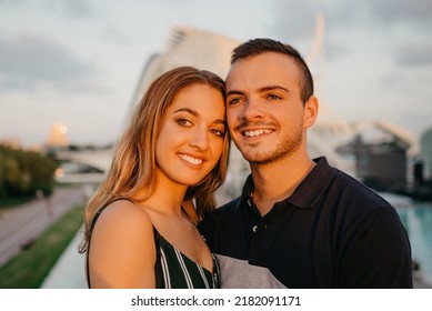 A Couple Of Smiling Tourists Are Having Fun With Modern Urban Space In The Background On A Date At The Sunset In Valencia. A Guy Is Hugging His Girlfriend In The Evening In Spain