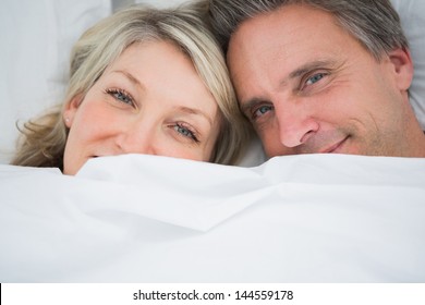 Couple Smiling From Under The Covers At Home In Bed