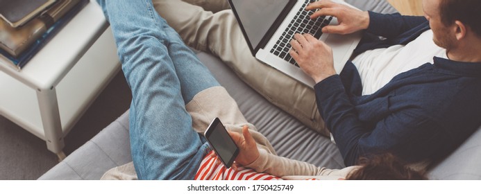 Couple Spending Time Together At Home. Man Using Computer And Woman Typing Message With Mobile Phone. Sitting Together On The Sofa. Modern Family Life. Wide Screen Panoramic