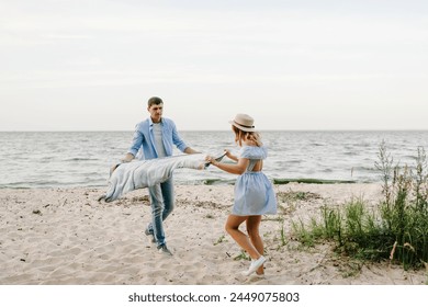 Couple spreading blanket for picnic on sand sea. Happy and funny couple in love on seashore. Female and male on beach ocean and enjoying summer day. Man and woman having fun spending time together. - Powered by Shutterstock