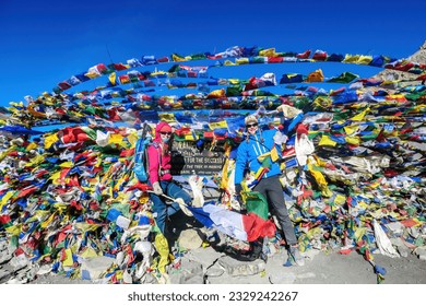 A couple standing between prayer flags at the top of Thorung La Pass, Annapurna Circuit Trek, Nepal. Memorial board. Colorful prayer flags attached to the stone wall, blown by the wind. Clear sky.