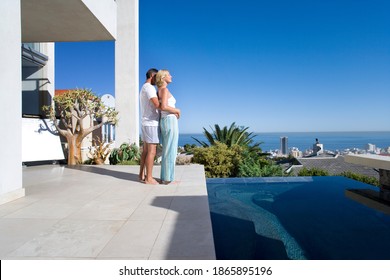 Couple standing and hugging on the balcony next to a swimming pool with the sea in the background