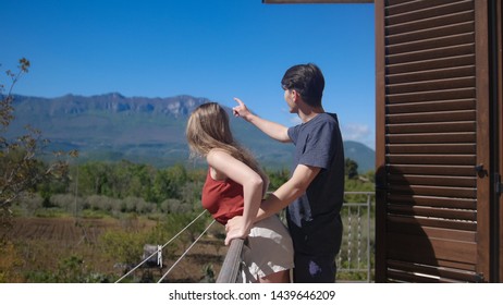 A Couple Is Standing On The Balcony Embracing. Look On The Mountain Landscape - Sideview