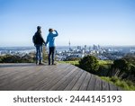 Couple standing on boardwalk at Mt Eden summit. Sky Tower and Auckland Harbour Bridge in the distance. Auckland.