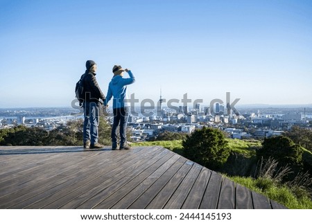 Couple standing on boardwalk at Mt Eden summit. Sky Tower and Auckland Harbour Bridge in the distance. Auckland.