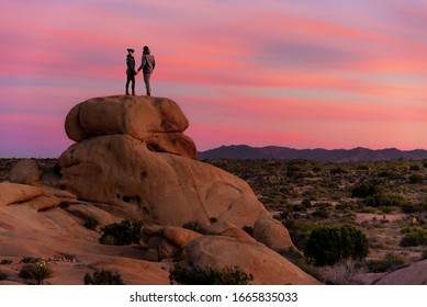 Couple Standing On A Rock In Joshua Tree National Park With Beautiful, Pink Sunset. 