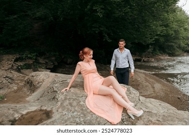 A couple is standing on a rocky shore, with a woman in a pink dress sitting on a rock - Powered by Shutterstock