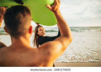 A couple is standing on a sandy beach with a green surfboard held above their heads. The man is facing away from the camera and the woman is looking back at the camera, smiling. - Powered by Shutterstock