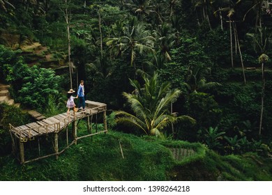 Couple standing on wooden bridge near rice terraces in Bali Indonesia. Holding hands. Romantic mood. Tropical vacation. Aerial shot. On background coconut palm trees. - Powered by Shutterstock