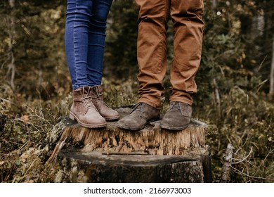 A Couple Is Standing On A Wooden Trunk And Their Boots And Jeans Are On The Photo. Close Up Shot Of Their Trousers And Brown Footwear. Outdoor Lifestyle In The Forest.	