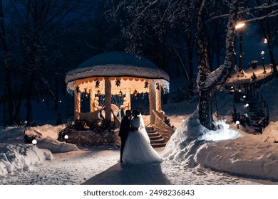 A couple is standing in the snow outside of a gazebo. The bride is wearing a white dress and the groom is wearing a suit. The scene is set in the winter and the couple is posing for a picture - Powered by Shutterstock