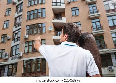 Couple Standing With Their Backs Towards Camera. Man Pointing To Apartment Window For Brunette Woman