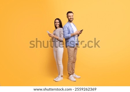 A couple stands back to back, smiling while using their smartphones. The cheerful atmosphere, highlighted by a vibrant yellow backdrop, captures their playful affection and connection.