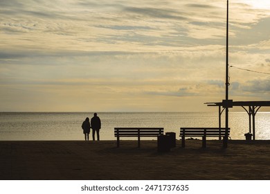 A couple stands by the seaside, silhouetted against a serene sunset. Empty benches and a lamppost frame the tranquil scene, with calm waters and a cloudy sky in the background - Powered by Shutterstock