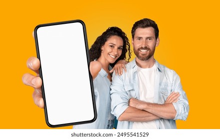 A couple stands in front of a bright yellow background. The woman has her arm around the man's shoulder and they both smile at the camera. The woman holds a smartphone with a blank screen - Powered by Shutterstock