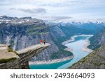 A couple stands on the edge of Trolltunga, Norway, a famous cliff in Norway, overlooking a breathtaking view of the surrounding mountains and a winding lake.