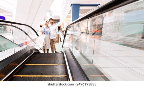 A couple stands on an escalator in a modern shopping mall, both smiling and holding shopping bags. The escalator is moving upward, with a clear glass side panel and a black metal handrail. - Powered by Shutterstock