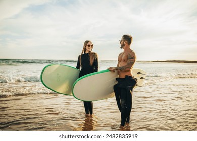 A couple stands on a sandy beach holding surfboards. They are both wearing wetsuits and sunglasses. The ocean is calm and the sky is clear. The sun is setting in the background - Powered by Shutterstock