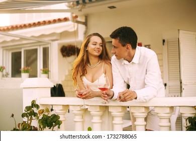 A Couple Stands On The Terrace And Smokes A Cigar, Drinks Red / Pink Wine. A Rich Man Is Resting In A House On The Ocean With His Girlfriend. Travel, Happiness, Laughter, Honeymoon. 