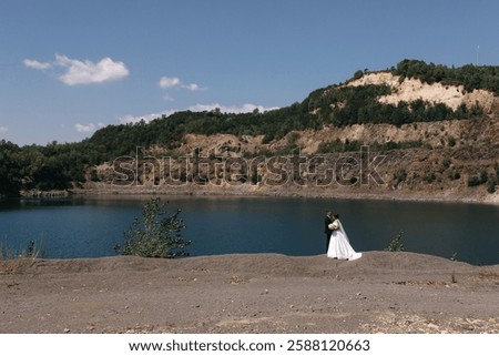 a couple stands with their backs to the camera on the shore of a pond, they are hugging, and in the background there is a blue lake
