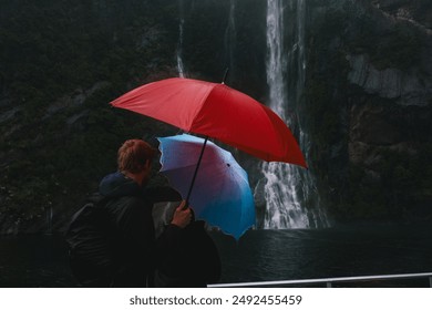 A couple stands under vibrant red and blue umbrellas, viewing a magnificent waterfall. The scene captures adventure and romance amidst a misty, lush landscape, with bright umbrellas contrasting with - Powered by Shutterstock