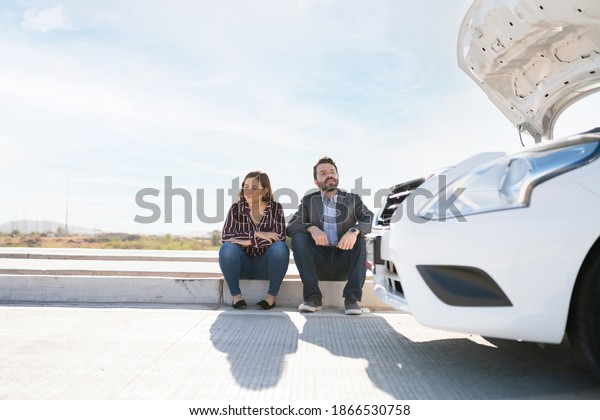 Couple stranded on the highway because their car
broke down. Young woman and adult man sitting and waiting for help
with the car hood open