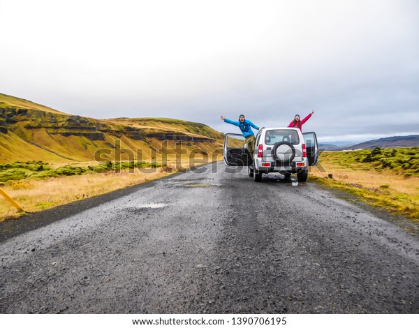 A couple stretching from the sides of the car and\
waving at the camera. The car is pulled on the side of the road,\
next to mountains. Mountains are overgrown with yellow and green\
grass. Fun moments
