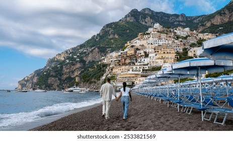 A couple strolls along a beach dotted with umbrellas, hand in hand, with vibrant cliffside villages perched above, as clouds gather over the breathtaking Amalfi Coast. - Powered by Shutterstock