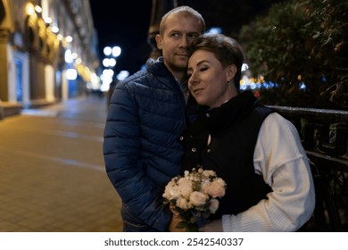 A couple strolls through the city at night, the man with his arm around his wife who holds a bouquet of flowers - Powered by Shutterstock