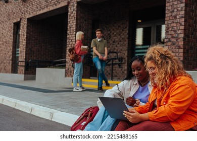 A Couple Of Students Are Working On A Laptop Outside University Building While A Couple Of Other Students Can Be Seen In The Background Having A Conversation. Modern And Stylish Campus Life.