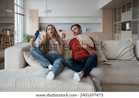 Couple suffering from hot sultry sweltering summertime weather at home. Sweaty exhausted wife and husband waving with hand fans sitting on sofa in overheated house in need of ventilator, conditioner