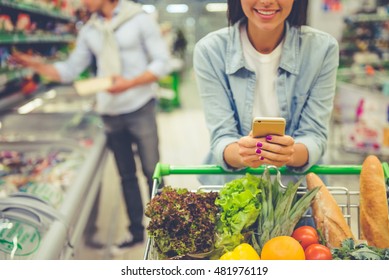 Couple In The Supermarket. Cropped Image Of Girl Leaning On Shopping Cart, Using A Mobile Phone And Smiling, In The Background Her Boyfriend Is Choosing Food