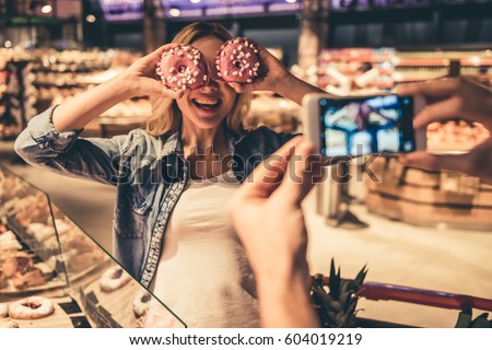 Couple at the supermarket. Girl is holding donuts and posing while her boyfriend is taking a photo
