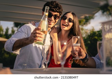 Couple of sweethearts standing on the table pouring sparkling wine champagne into their friends flute glasses celebrating the engaging party outdoors in the terrace garden - wearing sunglasses