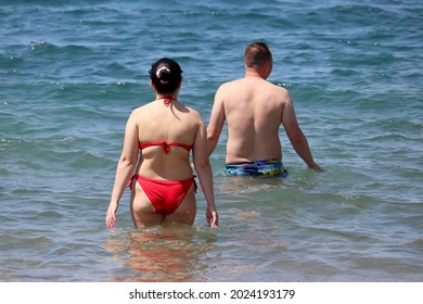 Couple In Swimwear Going To Swim In Sea Water, Rear View. Overweight Woman And Man On A Summer Beach, Family Leisure