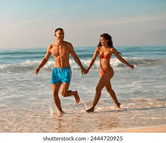 Couple In Swimwear On Vacation Holding Hands Running Along Beach Shoreline - Powered by Shutterstock