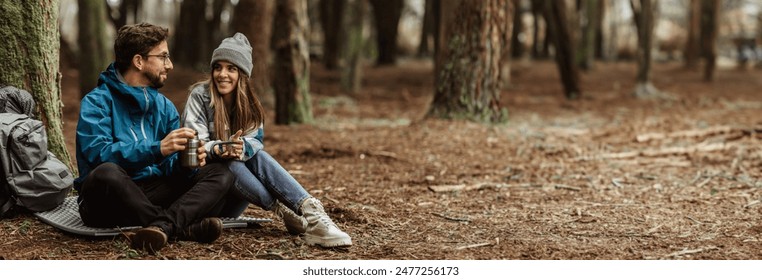 A couple takes a break from their forest hike to enjoy hot coffee in the serene forest setting. The man and woman are dressed in warm clothing, copy space - Powered by Shutterstock