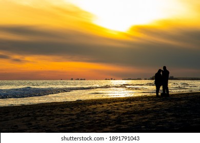 A Couple Takes A Selfie With A Phone On The Beach Near Sunset In A Coastal Travel Destination. The Ocean Reflects The Orange And Gold Light, Turning Them Into A Silhouette.