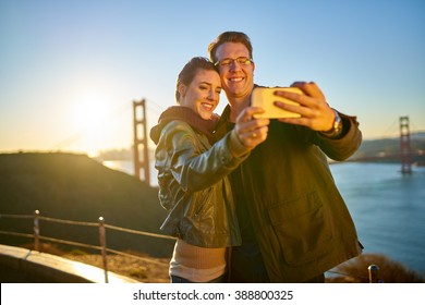 Couple Taking Selfie In Front Of Golden Gate Bridge With Sun Behind Them