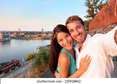 Couple taking selfie self portrait in Stockholm. Candid fresh Scandinavian man and Asian woman looking at old town cityscape sunset view from Monteliusvagen overlooking Gamla Stan, the old town.