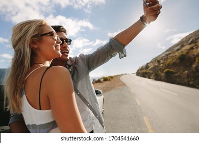 Couple taking a selfie using a smart phone and smiling while standing near car during travel. Man and woman posing for a self portrait by their car while on road trip.