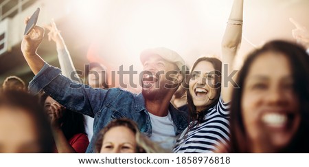 Couple taking selfie while cheering at stadium. Excited man and woman fans taking selfie while watching match in stadium.