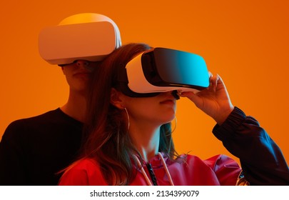 Couple Of Teen Boyfriend And Girlfriend Exploring Virtual Reality In VR Goggles On Orange Background In Studio With Neon Light